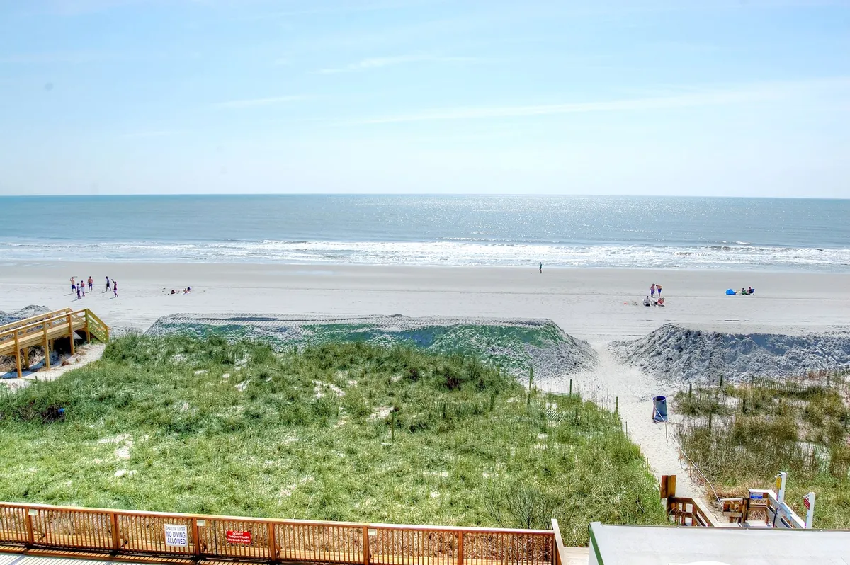 The photo captures a serene beach scene with the ocean, dune grass, a boardwalk, and people walking along the shore. The atmosphere is relaxed, suggesting sunset strolls and tranquil moments by the water.