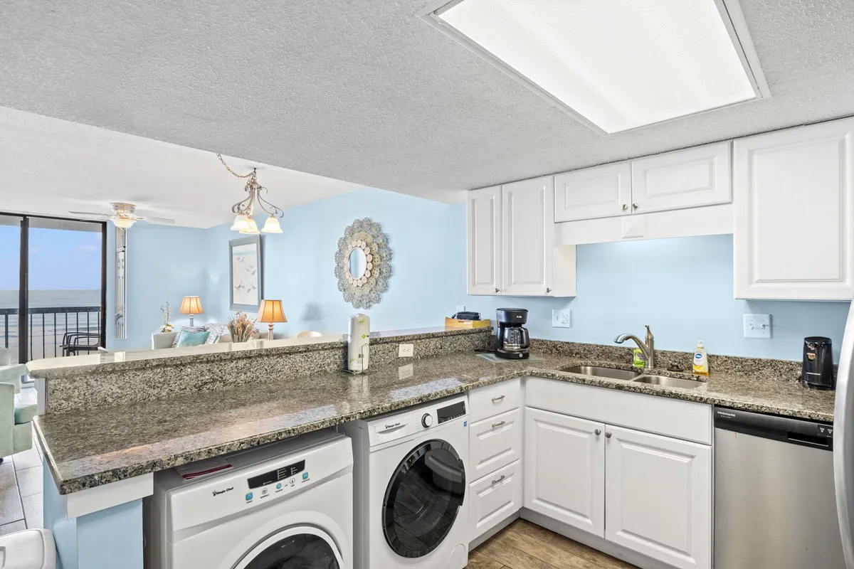 A kitchen scene with granite countertops, modern appliances, light blue walls, and a decorative mirror, all bathed in natural light from a window with a scenic view. The breakfast bar is visible, offering a spot to chat or enjoy a meal while taking in the landscape.