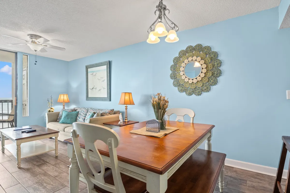 This view captures a cheerful interior with a dining table, a plush sofa, a decorative wall mirror, and coastal artwork. The space is designed for dining, relaxing, and socializing, with table lamps casting a warm glow, making it ideal for cozy gatherings.