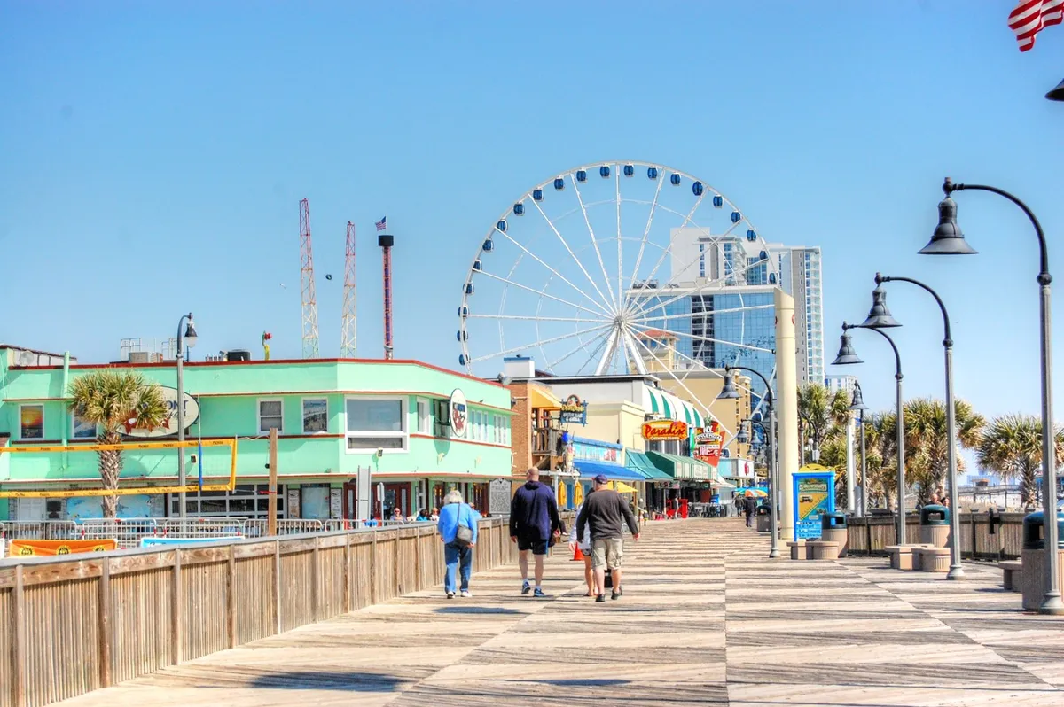 A lively boardwalk scene featuring a colorful Ferris wheel, vibrant storefronts, and families strolling along the promenade. The atmosphere is festive, with visible activity and people enjoying local attractions.