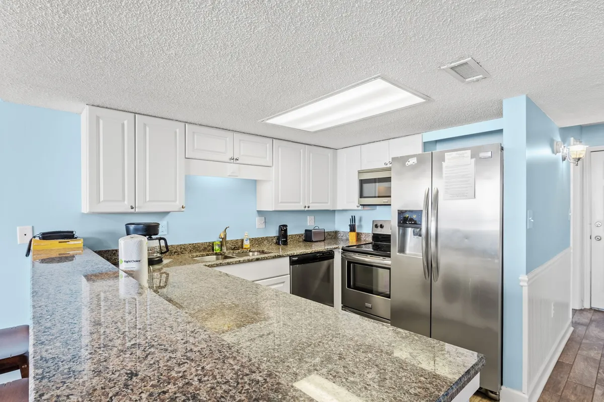 A contemporary kitchen angle showcasing spacious granite countertops, stainless steel appliances, a blue accent wall, and an open floor plan that encourages interaction. The image captures the energy of cooking, casual dining, and hosting friends or family.