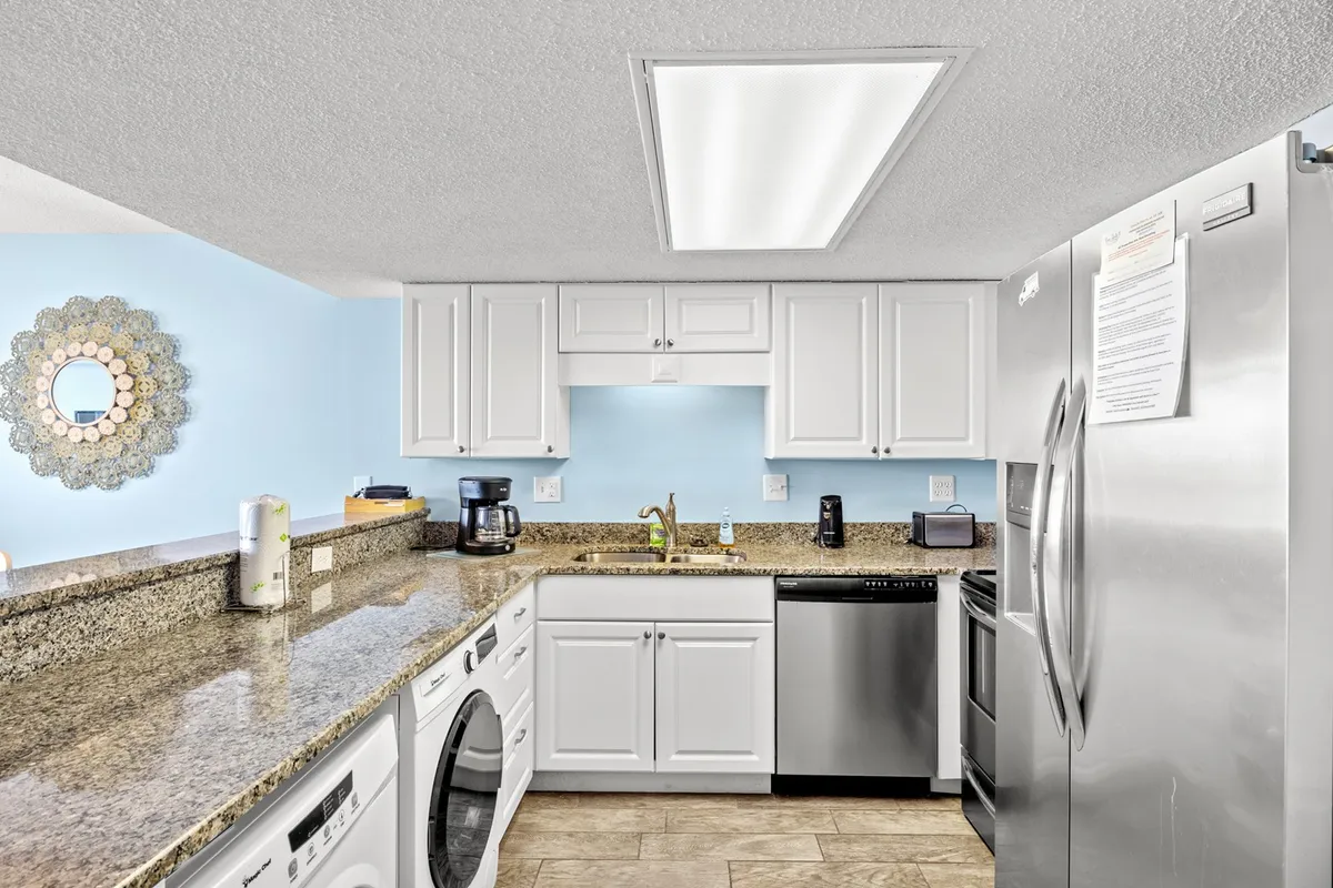 A bright, inviting kitchen featuring a granite countertop, gleaming stainless steel appliances, a light blue accent wall, and a decorative mirror. The open space shows plenty of room for food prep and gathering, with a breakfast bar area that connects the kitchen to the living space.