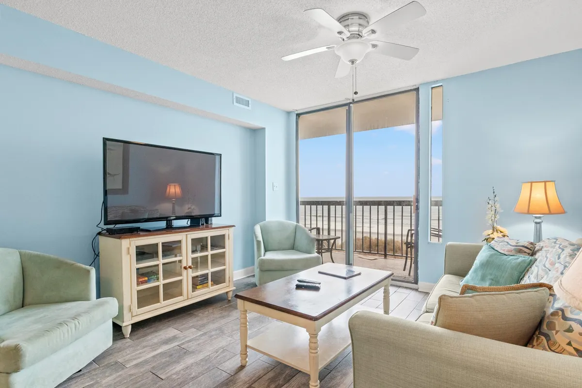 The image showcases a cozy, beautifully furnished living room with a comfortable seating arrangement centered around a coffee table and television. Large sliding glass doors open to a private balcony, revealing breathtaking ocean views—perfect for enjoying sunrises right from the comfort of the condo. The setup invites relaxation, socializing, and memorable family moments.