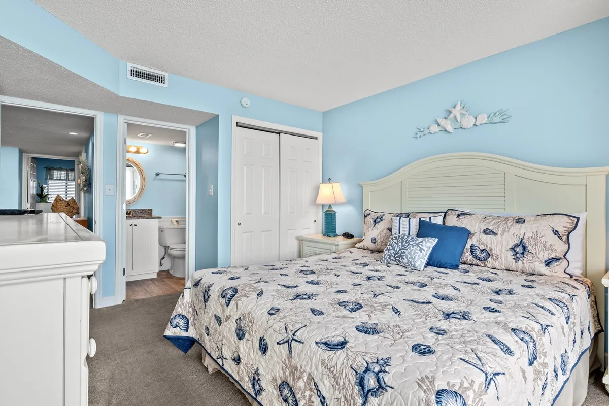 A cozy bedroom with seashell-patterned bedding, a bedside table with a lamp, and wall art featuring seashell motifs. Sunlight streams into the room, enhancing its serene and inviting mood.