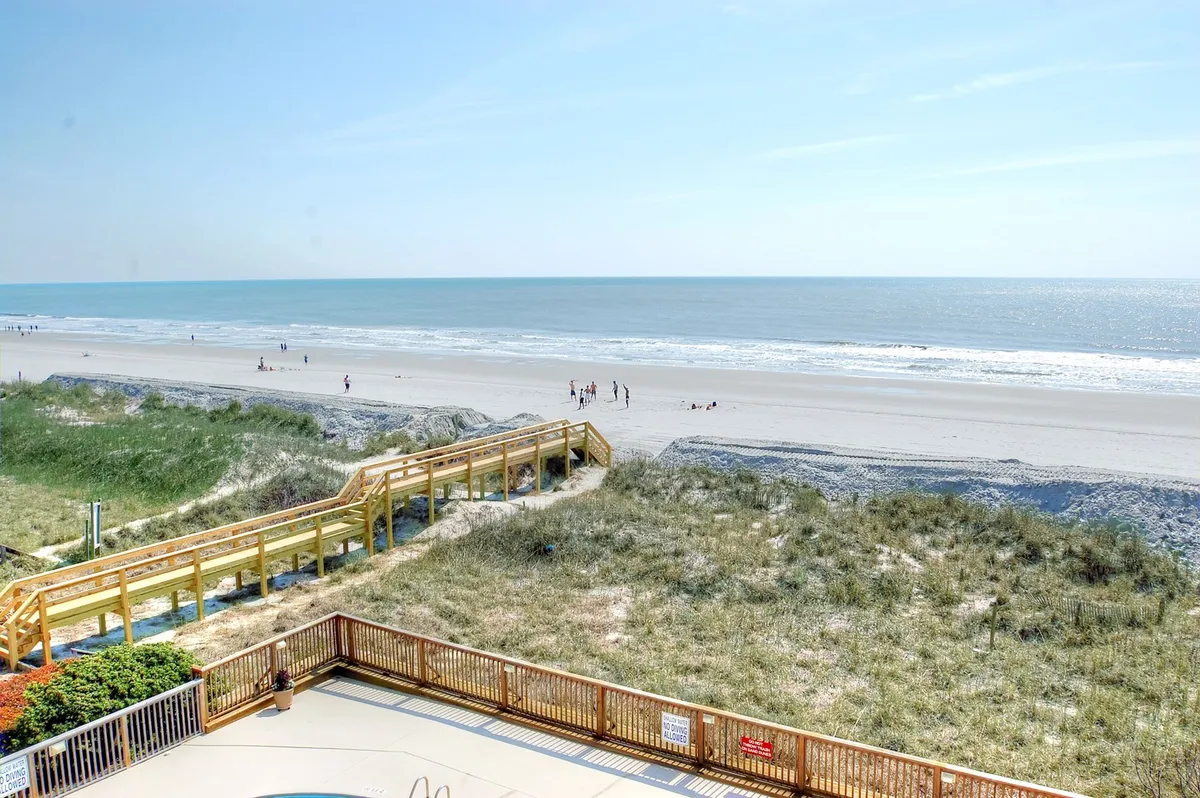 A serene beach scene featuring soft sand, a wooden walkway leading to the ocean, and families with children building sandcastles and enjoying the shoreline. Dune grass frames the view, connecting the property to the natural beauty of the coast.