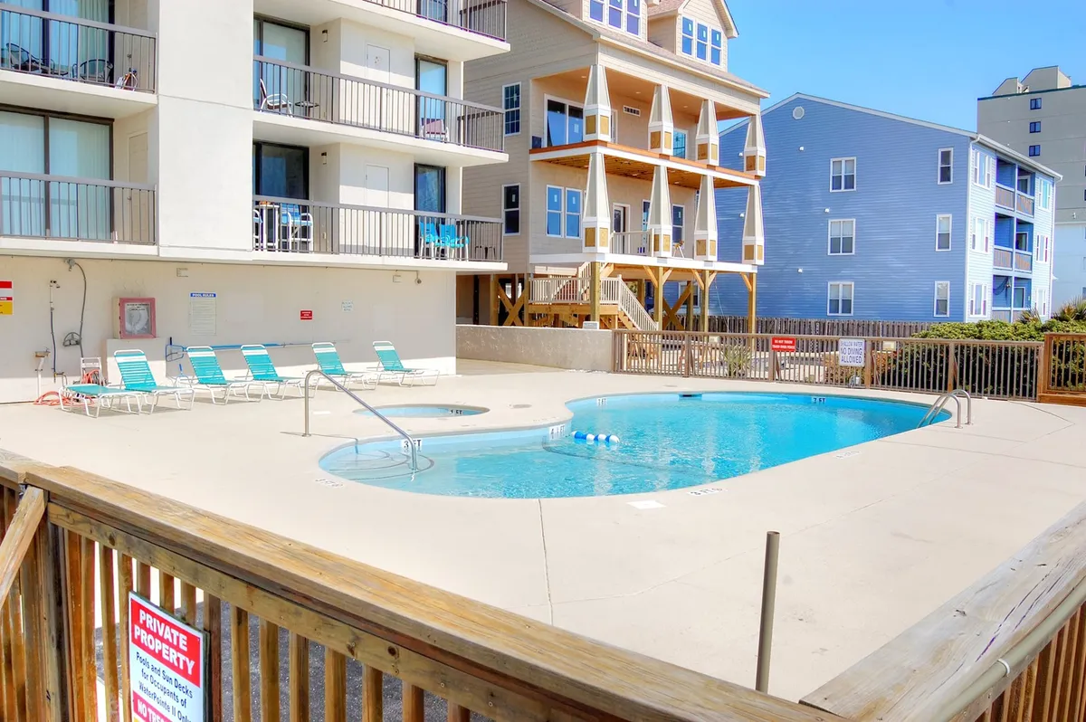An inviting outdoor swimming pool with lounge chairs arranged around it and building balconies visible in the background. The relaxed poolside atmosphere suggests a resort-style experience, ideal for both swimming and group sunbathing sessions.