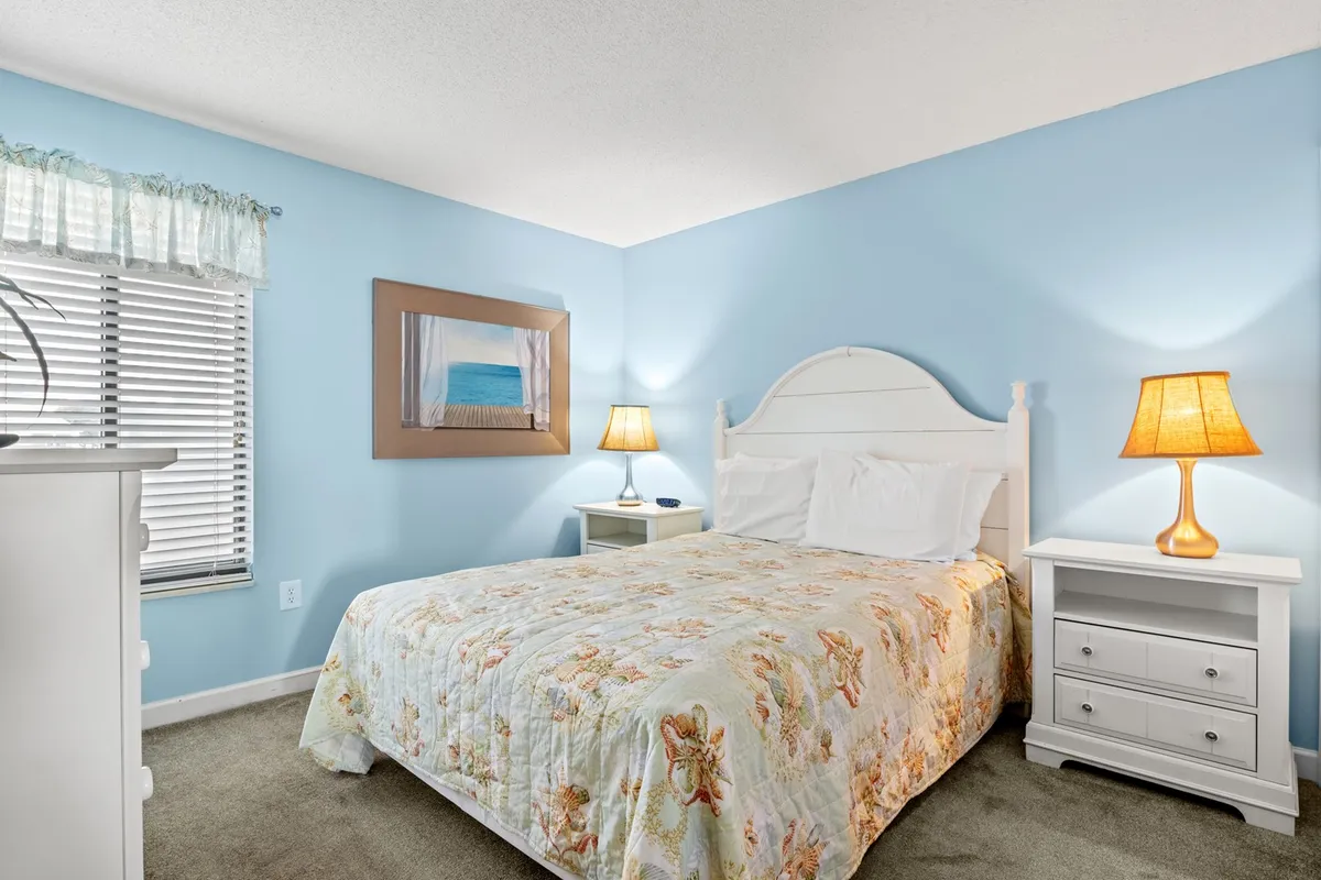 A bedroom showcasing a bed with a floral quilt, matching nightstands, framed beach artwork, and warm table lamps. The room is bathed in natural light, offering a relaxing environment ideal for unwinding after a day at the beach.