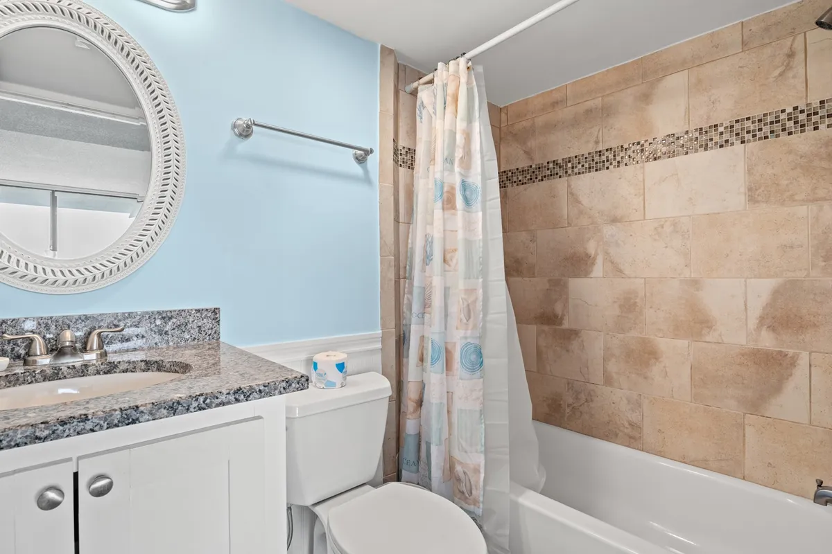 A serene interior shot of a bathroom with a granite countertop, framed mirror, and shower curtain, suggesting a comfortable and fresh place for cleanup after pool or beach activities.