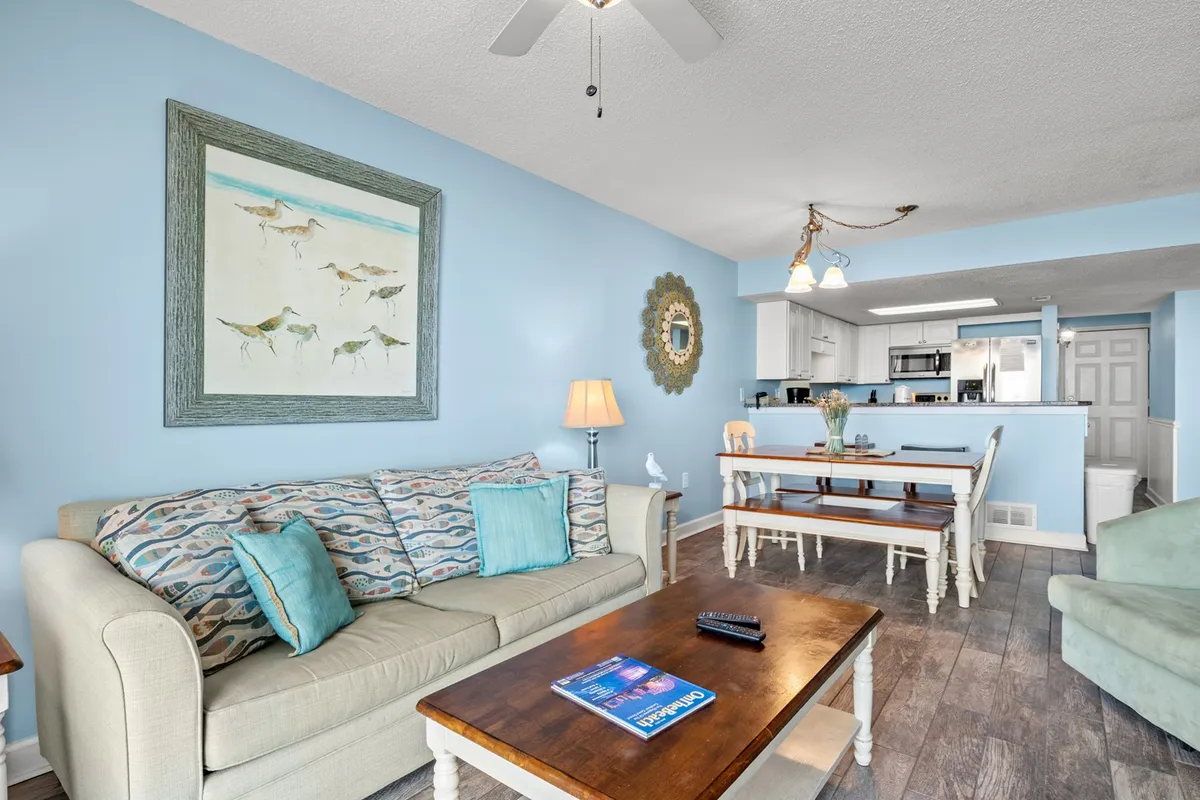 This image showcases a cozy and welcoming open-concept living and dining area. A sofa with patterned cushions sits adjacent to a dining table and kitchen area, with a painting of birds adding a touch of coastal flair. The space is bright and perfect for relaxing with family, entertaining guests, and enjoying group meals.