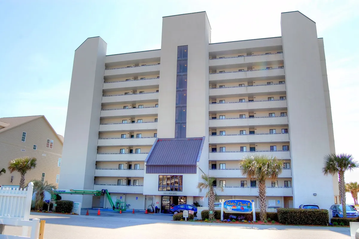 A wide exterior shot of a tall beachside condo building surrounded by palm trees, with the entrance area and signage visible. The scene conveys a welcoming, relaxed atmosphere perfect for long-term winter stays.