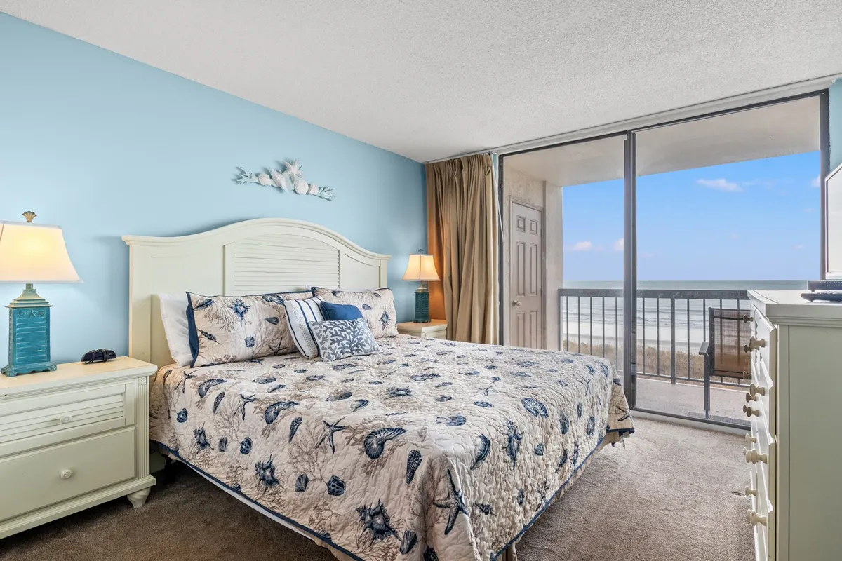 A tranquil bedroom featuring a bed with patterned bedding, flanked by a nightstand and lamp. Sliding glass doors open to a balcony, allowing natural light to fill the space and providing a glimpse of the outdoors, inviting guests to start their morning with views and fresh air.
