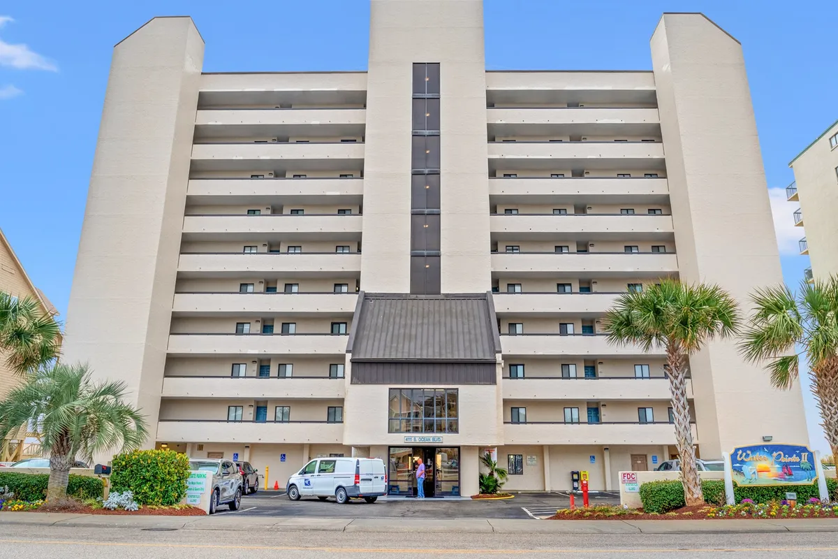 A welcoming exterior shot of the building’s main entrance, flanked by palm trees and featuring multiple balconies. The scene conveys a sense of arrival and hints at the communal, vacation-ready atmosphere of the property.